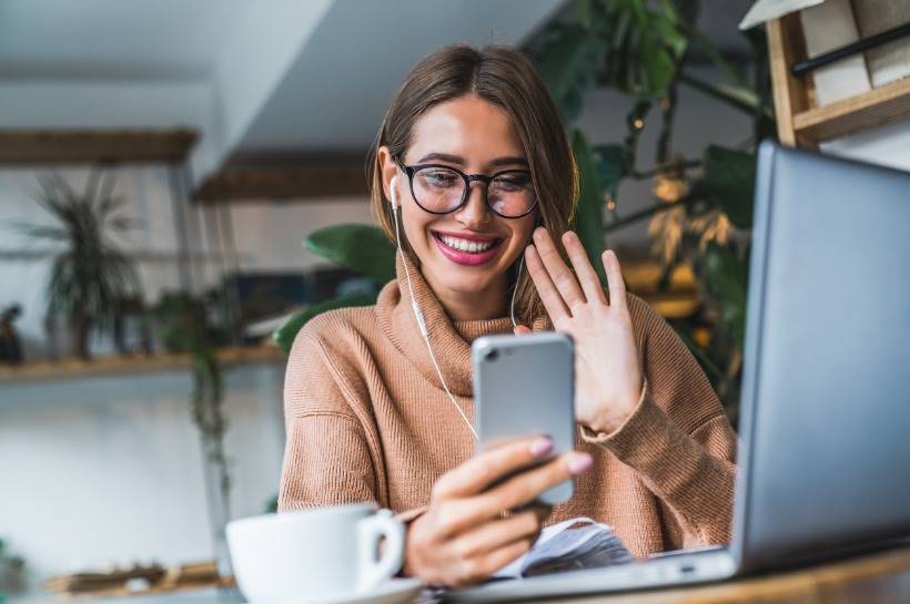 Woman wearing glasses, smiling and waving while video chatting.