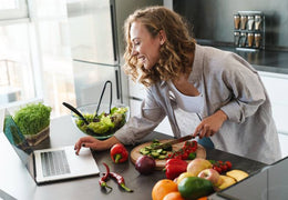 Woman smiling, looking at her laptop and chopping vegetables