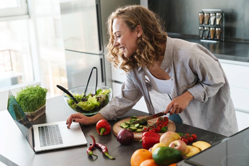 Woman smiling, looking at her laptop and chopping vegetables